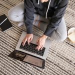 Woman in casual wear working remotely on a laptop, with a notebook and snack nearby.